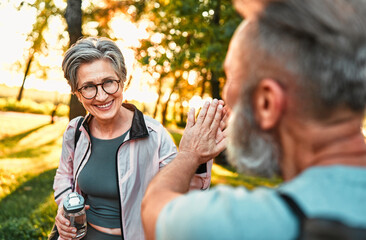Fototapeta premium Sincere intelligent modern beautiful smiling cheerful active stylish older lady in sportswear giving high five to friend after morning workout outdoors. Senior couple jogging together.