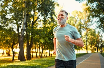 Tuinposter Concept of endurance. Well-shaped senior sportsman feeling joy when running in park. Cheerful male person wearing sport attire strengthening body muscles by every day jogging. © HBS