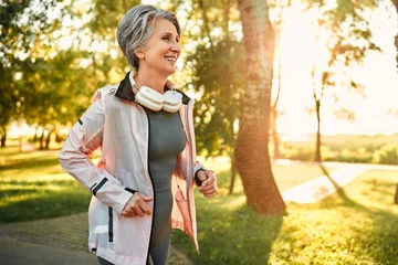 Muurstickers Remote technology and sport. Progressive aged lady with stylish grey haircut running in park with wireless headphones on neck. Slender female in sport outfit feeling contentment from morning jogging. © HBS