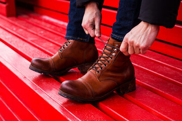 A man in jeans ties his shoelaces over brown shoes. Stylish men's shoes on a red wooden background - Powered by Adobe
