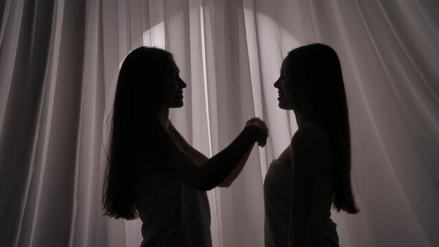 Medium slow motion video of two sisters, friends, young women's silhouettes wrapped in a towel. They are swishing each other's hair on a curtain background in a muffled light.