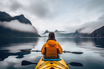 From the back view of a female tourist in a yellow kayak sailing along a sea bay with foggy fjords in the background.