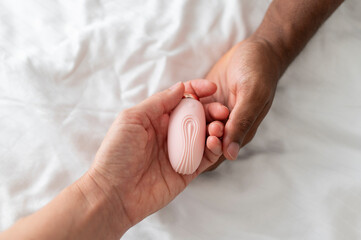Pink sex toy vibrator for mutual pleasure in hands of white woman and African man, on the white bedding - obrazy, fototapety, plakaty