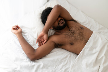Young African bearded man lying on the bed covering his face with arm from the sunlight, sleeping