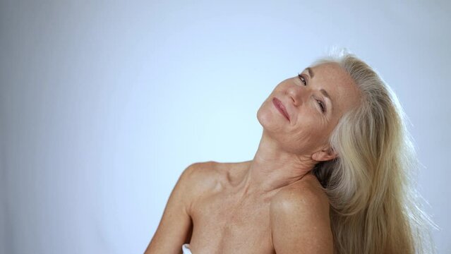 Slow motion portrait of smiling playful sexy flirty happy 50s, 60s middle aged woman with blond gray hair flipping it to side and putting hand on shoulder.