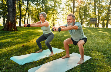Spouses working out with sport equipment. Angle view of athletic seniors doing squats with...
