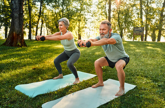 Spouses working out with sport equipment. Angle view of athletic seniors doing squats with outstretched arms while standing on fitness rugs. Determined couple training in park with dumbbells.