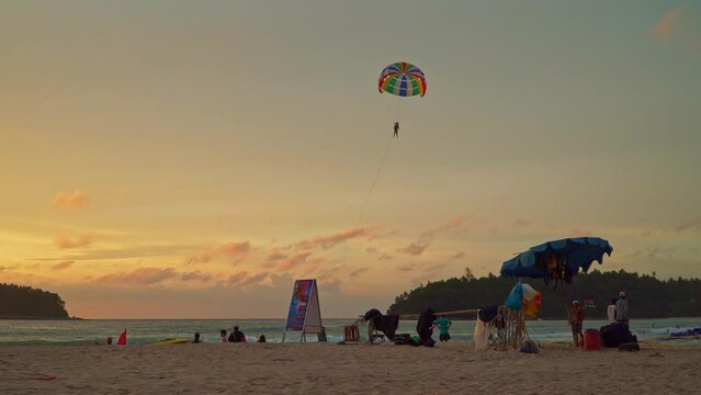 .Phuket,Thailand-August 12,2023: tourists relax on Kata beach Phuket..Tourists take photos on the beach under the golden sun..colorful parasailing in beautiful sunset..bright golden sky background.