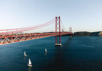 Aerial photo of the 25 April bridge (Ponte 25 de Abril) located in Lisbon, Portugal, crossing the Targus river - taken by drone. The 25 April bridge with sailing boats under the bridge on blue water