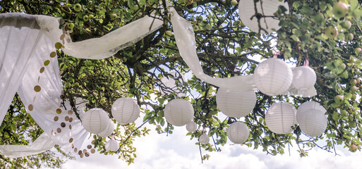 viele weiße Lampions, Papierlaternen, Glitzerkette und weißes Tuch dekorativ, festlich hängen im Apfelbaum. Für Festival, Party, Hochzeit