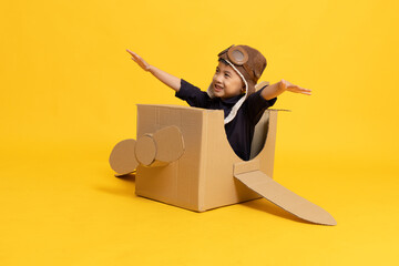Asian little boy aviator sitting and playing with cardboard airplane isolated on yellow background