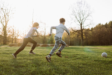 Rear view of soccer players purposefully running towards ball, playing football on lawn at sunset and enjoying game.