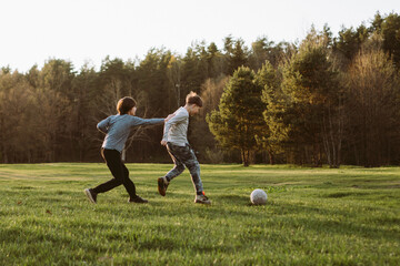 Foxy teenager grabbing friend on clothes attempting to overtake boy running towards ball, playing football in nature. - obrazy, fototapety, plakaty