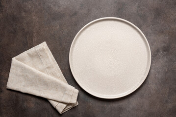 Empty beige plate with beige linen napkin on a dark brown background. Top view, flat lay.