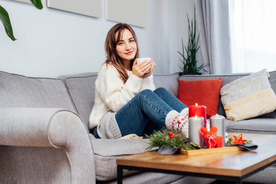 Portrait of smiling woman with cup of tea on sofa at home with christmas decoration atmosphere. Lady wear jumper and warm socks. Cozy and comfortable winter concept. Selective focus. Copy space.
