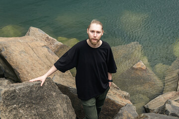 A young man with a beard and a bun in a gray T-shirt stands among the stones near the lake or the...