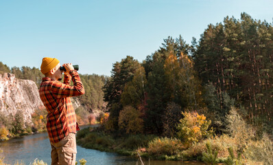 young man in red plaid shirt and yellow beanie hat looking through binoculars on river bank near cliff in fall Birdwatching and tourism