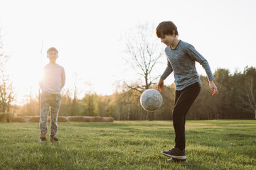 Glorious boy kicking soccer ball up, performing skills on lawn while smiling friend standing near and watching process.