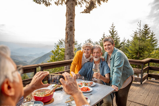 Senior group of friends having their picture taken on smartphone while having lunch and wine on a balcony of a cabin house in the forest