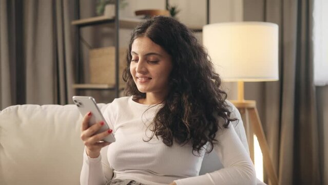 Charming young curly woman hold smartphone scrolling app watching social media feed and looking at the screen indoors Happy female texting enjoying leisure time laying on couch at cozy home