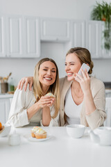 Obraz na płótnie Canvas lovely laughing sisters tasting cupcakes and holding phone on kitchen background, family bonding