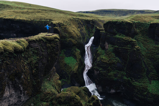 A man on a cliff is feeling free and enjoying nature in Iceland. There is a waterfall.