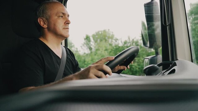 A man keeps his hands on the steering wheel and drives a truck down the road on a warm summer day. The truck driver is driving to the destination. View from the cab of the truck.