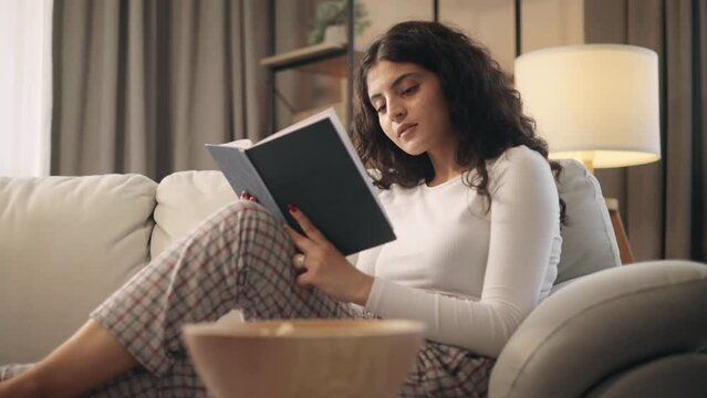 Portrait of charming curly young woman laying on sofa and reading book literature at home Calm relaxed female enjoying weekend leisure time alone indoors. High quality 4k footage