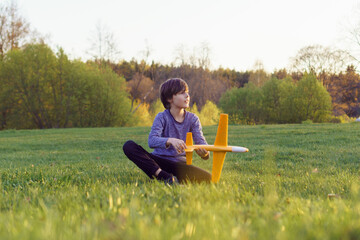 Delightful teenage boy sitting on lawn, holding plane toy and looking away, mesmerizing sunset and enjoying leisure.