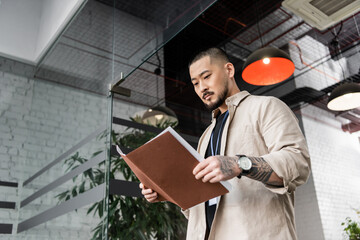 focused asian team leader with tattoo looking at startup plan inside of folder near glass door