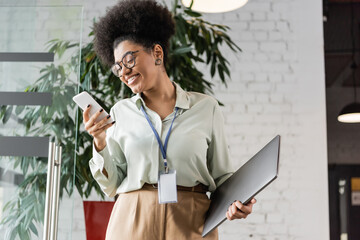 positive african american businesswoman in glasses holding laptop and using smartphone in office