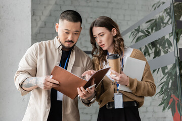 pretty businesswoman pointing at folder and discussing startup project with focused asian colleague