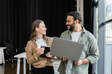happy indian businessman with laptop explaining task to female coworker with notebook, mentorship