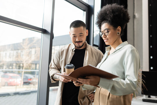 african american woman showing startup project in folder to asian businessman in coworking