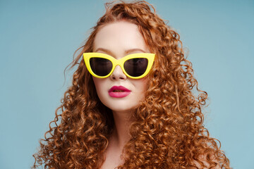 Closeup portrait beautiful stylish redhead woman wearing yellow sunglasses isolated on blue background