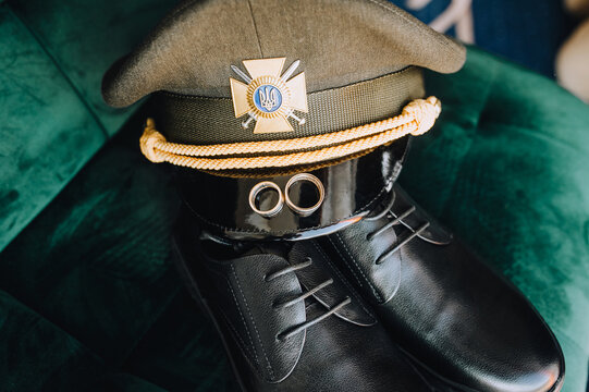 Gold rings, a cap, leather shoes, the uniform of a Ukrainian military groom on a chair. Wedding photography, accessories, details. War in Ukraine.