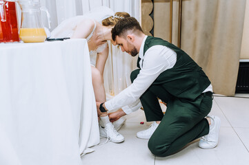 A stylish young groom in a suit helps a beautiful bride in a white dress put on shoes, change shoes while sitting at a table in a restaurant at a table. Wedding photography, portrait.
