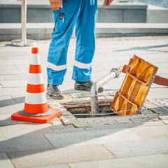 Worker standing next to open sewer manhole near traffic cone. Repair of sewerage and underground...