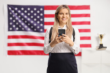 Happy female student holding a smartphone and typing in front of USA flag