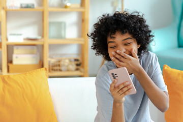 Fototapeta na wymiar Excited happy young black african american woman holding cell phone laughing feeling joy getting mobile message. Overjoyed girl laughing aloud sitting on couch watching funny video reading news