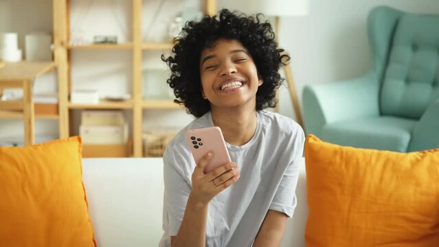 Excited Happy Young Black African American Woman Holding Cell Phone Laughing Feeling Joy Getting Mobile Message. Overjoyed Girl Laughing Aloud Sitting On Couch Watching Funny Video Reading News