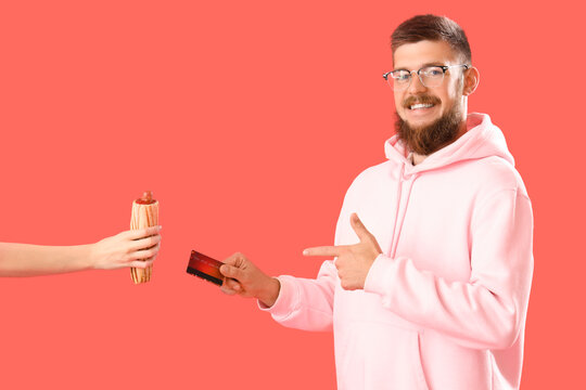 Portrait of handsome young man buying tasty hot dog with credit card on red background - Powered by Adobe