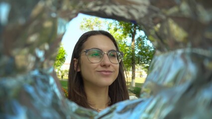 A young woman with glasses eats potato chips from a pack while walking in the park. View of a woman from a bag of chips.