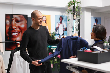 Clothing store asian woman employee consulting arab man buyer in choosing formal wear. Shopping mall assistant and customer holding jacket on hanger and discussing garment style