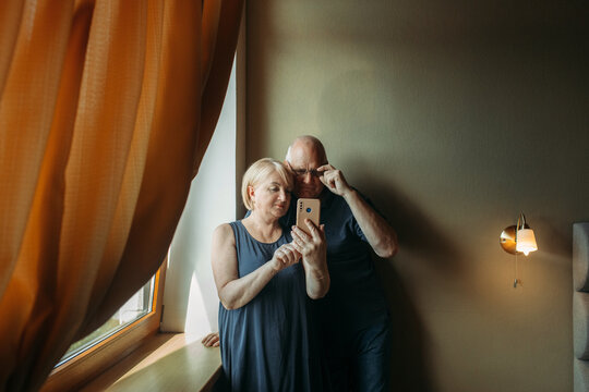 A happy elderly couple stand near the window, pose, take a selfie on the phone