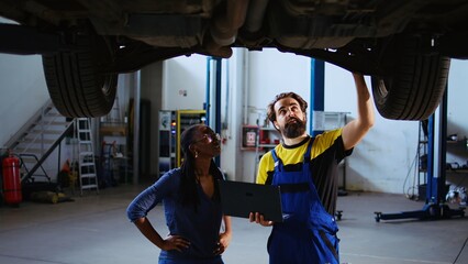 Repairman and customer standing underneath car in garage using laptop to order new parts after finding issues. Worker assisting client by looking online for replacing vehicle components