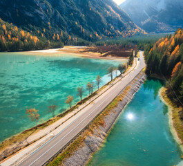 Aerial view of road, azure lakes, forest in alpine mountains in autumn sunny day. Dolomites, Italy. Travel. Top view of beautiful road, blue water, orange trees in fall. Landscape with highway. Nature