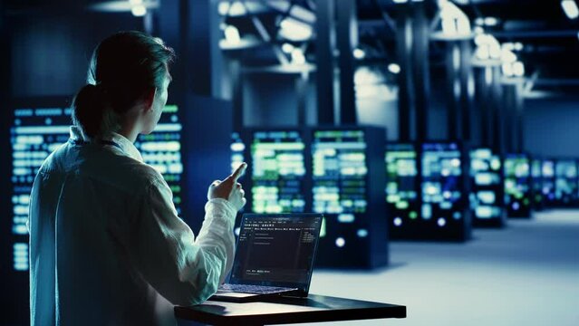 Asian system administrator running code and programming on laptop, troubleshooting and upgrading server farm equipment designed to accommodate mainframes, networking racks and storage arrays