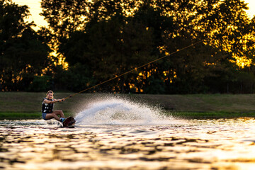 Female wakeboarder wakesurfing on sunset. Girl riding waterski cabel. Holding tow rope. Summer...