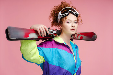 Beautiful curly haired woman, skier wearing stylish overalls, ski googles, holding ski equipment looking at camera isolated on  background. Fashion model posing in studio. Travel, vacation, ski resort - obrazy, fototapety, plakaty